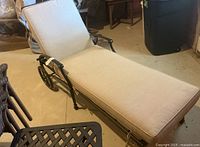 Full view of black metal chaise lounge with beige cushions laying flat indoors on concrete floor, showing wheels on one end for mobility.