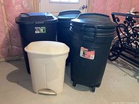 Four plastic garbage cans arranged on basement floor: two green Rubbermaid Roughneck 32 gallon cans on wheels, one green Ecoark 45 gallon can on wheels, and one smaller beige pedal can without wheels.