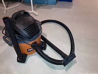 Orange and black Ridgid shop vac angled view with flexible hose and floor nozzle attached, placed on floor.