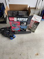 Toro Super electric leaf blower vac with box, blower tube, and hearing protection earmuffs on concrete floor near garage door.