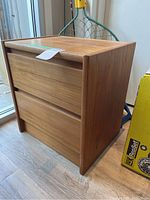 Side angle of the wooden bedside table showing its natural wood finish and two drawers.