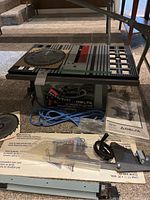 Front view of the Delta 10" Deluxe Bench Saw with two blades on table surface, instruction manual and plastic blade guard in front, blue power cable coiled underneath.