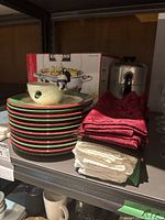 Shelf display showing stacked Christmas-themed plates with colorful rims and holiday motifs, including the Kirkland boxed chafing dish, metal percolator, and stacked cloth napkins in red and white.