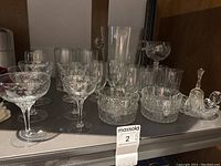 Front view photo showing a variety of crystal glassware including coupes, wine glasses with floral etching, small bowls, an ice bucket, and decorative figurines on a shelf.