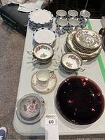 Overview of assorted cups, saucers, and plates on table showing different patterns and colors including floral, geometric, and solid dark red plate.