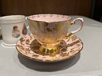 Pink cup with gold intricate patterns on matching saucer, showing ornate design and detailing.