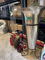 Photo showing large hammered metal umbrella stand on black metal tripod, tall clear acrylic vase, small brown pot with red berries, and multiple red glass candle holders including black metal candle holder.