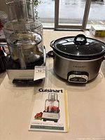 Photo showing both the Cuisinart food processor and Crockpot slow cooker with instruction booklet for the food processor in front, placed on a table near windows.
