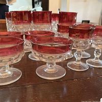 Photo showing 10 pieces of glassware with clear bases and ruby red tinted tops arranged on a wooden surface. Includes glasses and dishes of various heights and shapes.