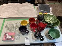 Full view of assorted housewares on table including cups, mugs, cookie cutter, green leaf bowls and plate, BMW travel mug, and two clear textured glasses.