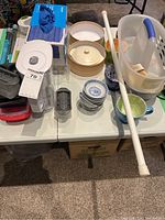 Full assortment of kitchen lot items displayed on table including cups, ceramic brie baker, microfiber cloths, tension rods, plastic containers and baskets.