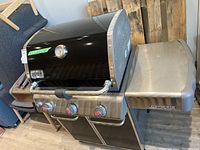Photo showing the full outdoor grill with black hood, three burner knobs, side shelf, and cabinet underneath. Grill is indoors on floor.