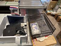 Overview of the lot showing a white plastic bin containing a black keyboard with blue cover, metal mesh file holders, boxes, and various cables. Large translucent plastic storage box with gray lid seen behind the bin, also visible is packaging for the door knob.