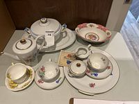 Porcelain serving set on a white table showing coffee pot, cups, saucers, covered serving platters, sugar bowl, creamer, and assorted plates.