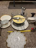 Wide view showing majority of items including cake cutter, cake plate, deviled egg platter, marble lazy Susan, salt and pepper shakers, 5-piece crème brûlée set wrapped, Pyrex gravy bowl, and kitchen torch arranged on carpet with measuring tape for scale.
