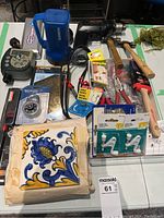 Overview of lot with various hand tools including hammers, pliers, wrenches, power drill, flashlight, packaged ceramic tile, hooks, drill bits, lock and paintbrush.