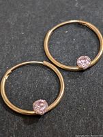Close up of two small 10K gold hoop earrings each with one pink cubic zirconia stone on black background.