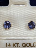 Pair of 14K gold stud earrings with round blue-violet iolite stones mounted in yellow gold prong settings displayed on cream textured card showing 14 KT GOLD mark.