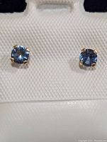 Close-up of pair of 14K gold stud earrings with blue-violet round tanzanite gemstones held by four prongs on a white textured display card.