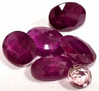 Close-up of six loose faceted gemstones placed on white surface. Five are purple-red rubies and one is a smaller pink gemstone.