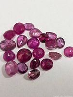 Close-up image showing a collection of 21 loose faceted ruby gemstones in varying shapes and shades of red to pink.