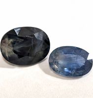 Close-up of two oval cut sapphire gemstones, one dark and one lighter blue, showing facets and internal inclusions.