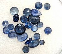 Close-up photo of multiple loose blue sapphire gemstones showing various cuts and shades of blue on a white background.