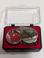 Jeweler's loupe open inside black plastic case with clear hinged lid, red velvet base