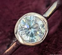 Close-up of the round, brilliant-cut moissanite gemstone set in a silver bezel setting on a thin silver band against a dark velvet background.