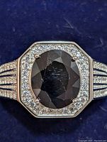 Close-up front view of the silver rhodium plated ring showing an oval faceted dark blue sapphire center stone surrounded by small clear stones and detailed multi-row band with stones.