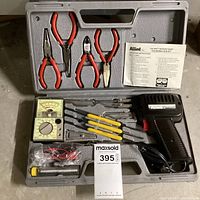 Photo showing open gray carrying case with Allied soldering iron gun, six yellow-handled soldering tips, analog multimeter, four red-handled pliers, test leads, metal file, and instruction manual inside.