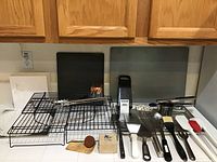 Photo showing cooling racks, baking sheets, metal graters, and various spatulas and utensils in black, metal, and wooden finishes.
