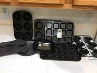 Full view of multiple baking pans including muffin pans, Bundt pans, tart pan, rectangular pan, and oven mitts laid out on countertop.