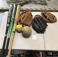 Photo of three bats, two balls, and three gloves on white folding tables.