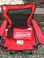 Red Coleman Broadband mesh quad camping chair unfolded outside on gravel showing seat, mesh back, armrests with cup holders and bundled second chair in front