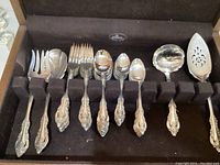Overview of flatware pieces arranged in velvet-lined wooden box showing spoons, forks, cake slicer, and gravy server with detailed ornate handles.