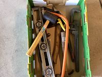 Photo showing a box with a wooden-handled hammer, black rubber mallet, orange hacksaw, pipe wrench, and metal spirit level.