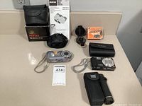 Full view of all items on countertop including both digital cameras, binoculars, their cases, and the Lensatic compass with box.