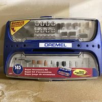 Front view of the Dremel 145-piece Super Accessory Kit in a blue case showing various rotary tool bits and accessories including grinding stones, sanding drums, and polishing wheels. Label highlights bonus sharpener.
