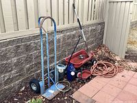 Blue metal dolly, coiled garden hose, water cans, small red wagon, and other garden tools