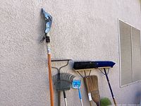 Photo showing tree limb cutter with orange handle, green rake, blue push broom, corn broom, push broom with blue head, green duster brush leaning against wall