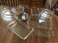 Various sized clear glass baking pans on a wooden table, including pie pans, rectangular loaf pans, and square baking dish.