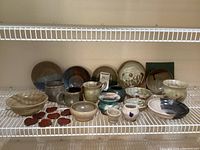 Photo showing the assortment of handmade pottery items including bowls, mugs, plates, and small decorative heart pieces arranged on a shelf.