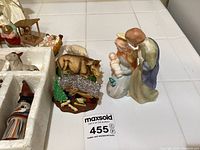 Top view of nativity scene figurines including Mary, Joseph holding baby Jesus, a cow, nativity stable and other animal figurines on a white tiled surface.