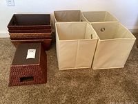 Overview of six woven baskets in various shades of brown stacked and arranged with four beige canvas folding containers nearby on carpeted floor.