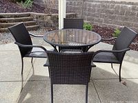 Full set showing four brown woven chairs around a round table with tinted glass top outdoors on concrete.