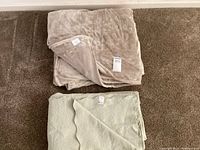 Two blankets folded and laid on carpeted floor, tan plush Charisma blanket on top, light green cotton bedcover underneath.