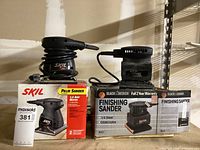 Two electric sanders on top of their original packaging boxes, one identified as SKIL palm sander and the other Black & Decker finishing sander.