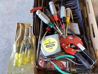 Photo of assorted hand tools including drivers with colored handles, red clamp tool, Stanley tape measure, pliers, and screwdriver bits inside a box