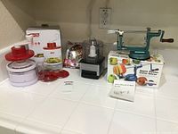 Wide view of all three kitchen appliances on white counter: green apple peeler corer slicer, DASH spiral slicer in box, CUISINART chopper